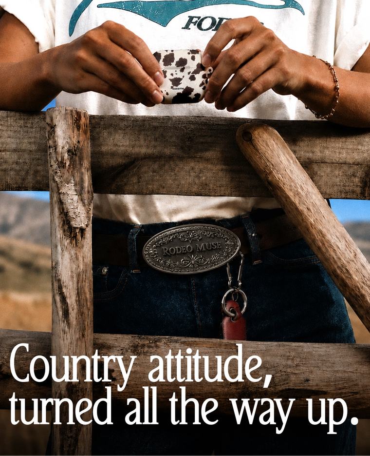 A person outdoors behind a rustic wooden fence, wearing a white graphic T-shirt, dark jeans, and a large engraved silver belt buckle that reads 'RODEO MUSE.' The person is holding a small cow-print item between their hands.
