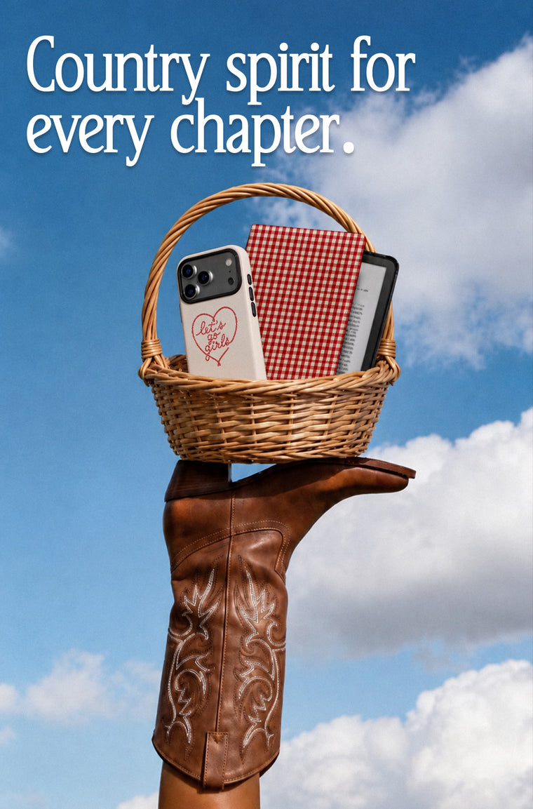 A hand wearing a brown western-style cowboy boot holds a wicker basket containing a phone case with a red heart design, a red-and-white gingham notebook, and a phone case against a blue sky with clouds. White text at the top reads, 'Country spirit for every chapter.