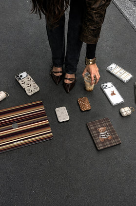 A top-down view of multiple designer accessories with brown and beige stripes, including a laptop skin, AirPods case, phone cases, and a ChargePods, arranged on a dark gray floor with a person's legs and hand visible holding a drink.