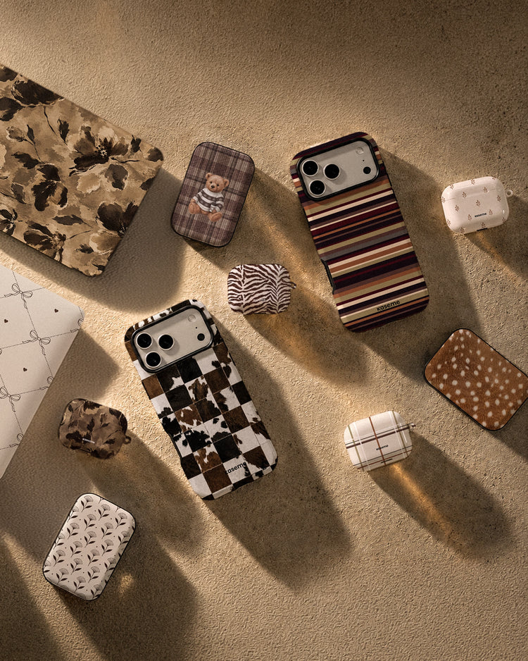 A styled flat-lay of multiple phone cases arranged on a textured beige surface with warm directional sunlight and shadows. The cases feature various patterns including stripes, plaid, checkered motifs, zebra print, polka dots, and floral designs in earthy tones and white.