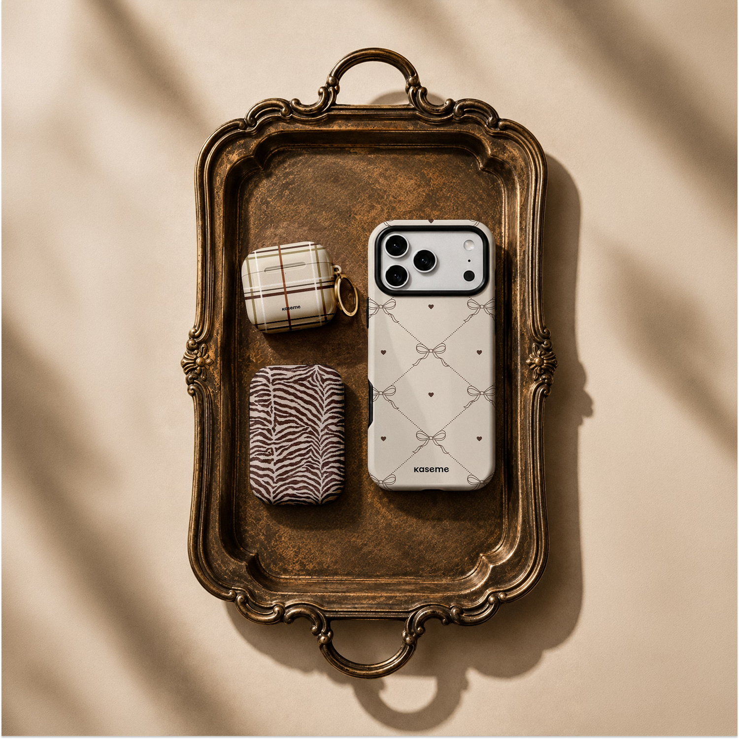 A white phone case with a bow and diamond pattern sits on an ornate vintage-style metal tray, styled with two small pouches. The scene is shot from a top-down angle on a beige background with soft diagonal shadows.