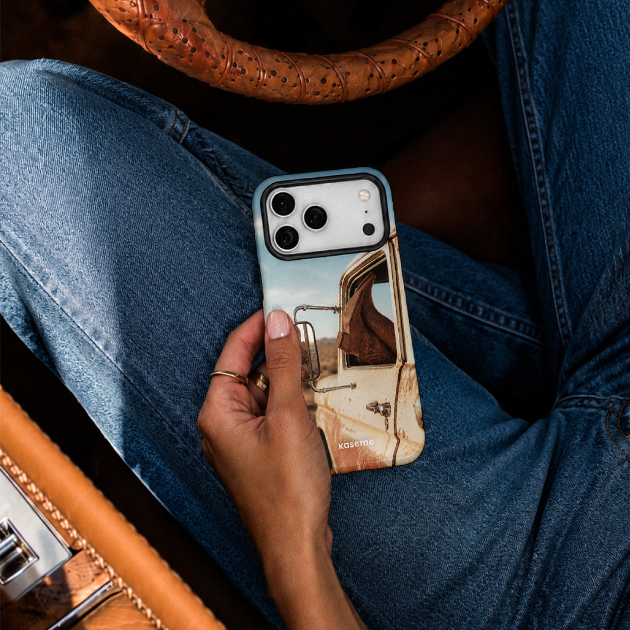A person in blue jeans holds a smartphone with a printed case featuring a vintage truck illustration. The scene is styled with a tan leather steering wheel and interior visible in the background.