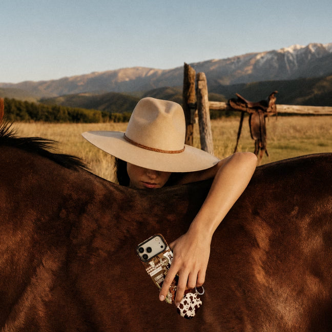 A person in a wide-brimmed beige hat leans over a brown horse in an outdoor ranch setting, holding a patterned phone case near the horse's neck, with mountains and a wooden fence in the background.