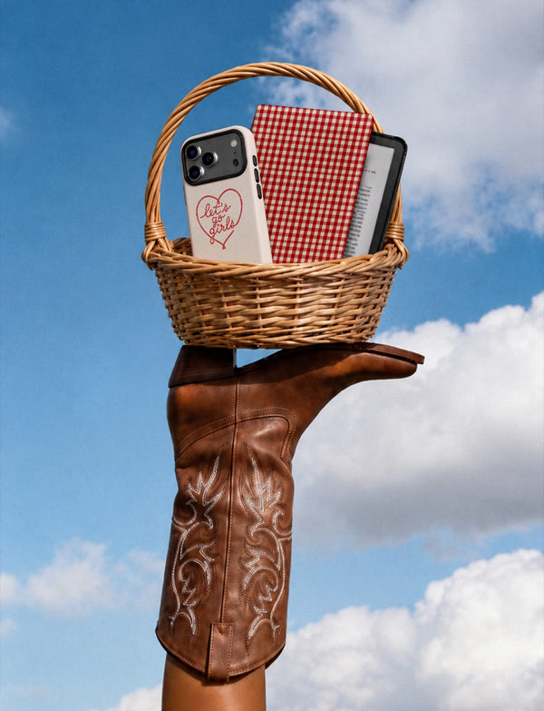 A hand wearing a brown leather cowboy-style glove holds a small wicker basket against a blue sky with clouds. Inside the basket are a white phone case with a red heart and 'Let's go girls' script, a red-and-white gingham notebook or book, and a black smartphone.